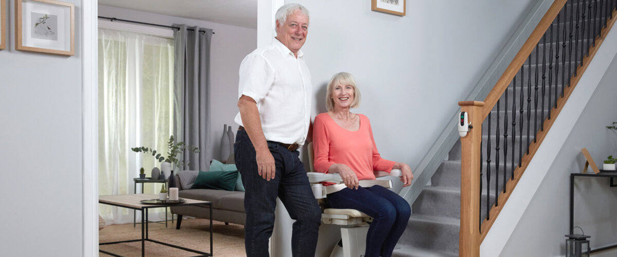 A seated woman and standing man smile near a stairlift inside a home.
