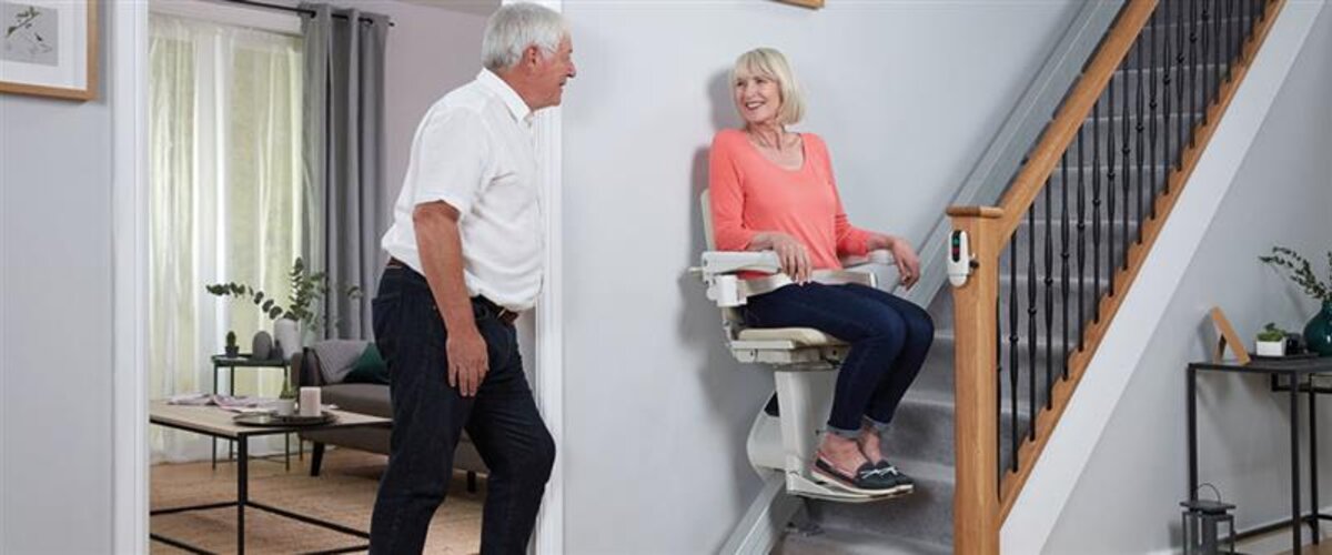 A stairlift is ascending a carpeted staircase; a woman is seated in it, smiling at a man standing at the bottom of the stairs.