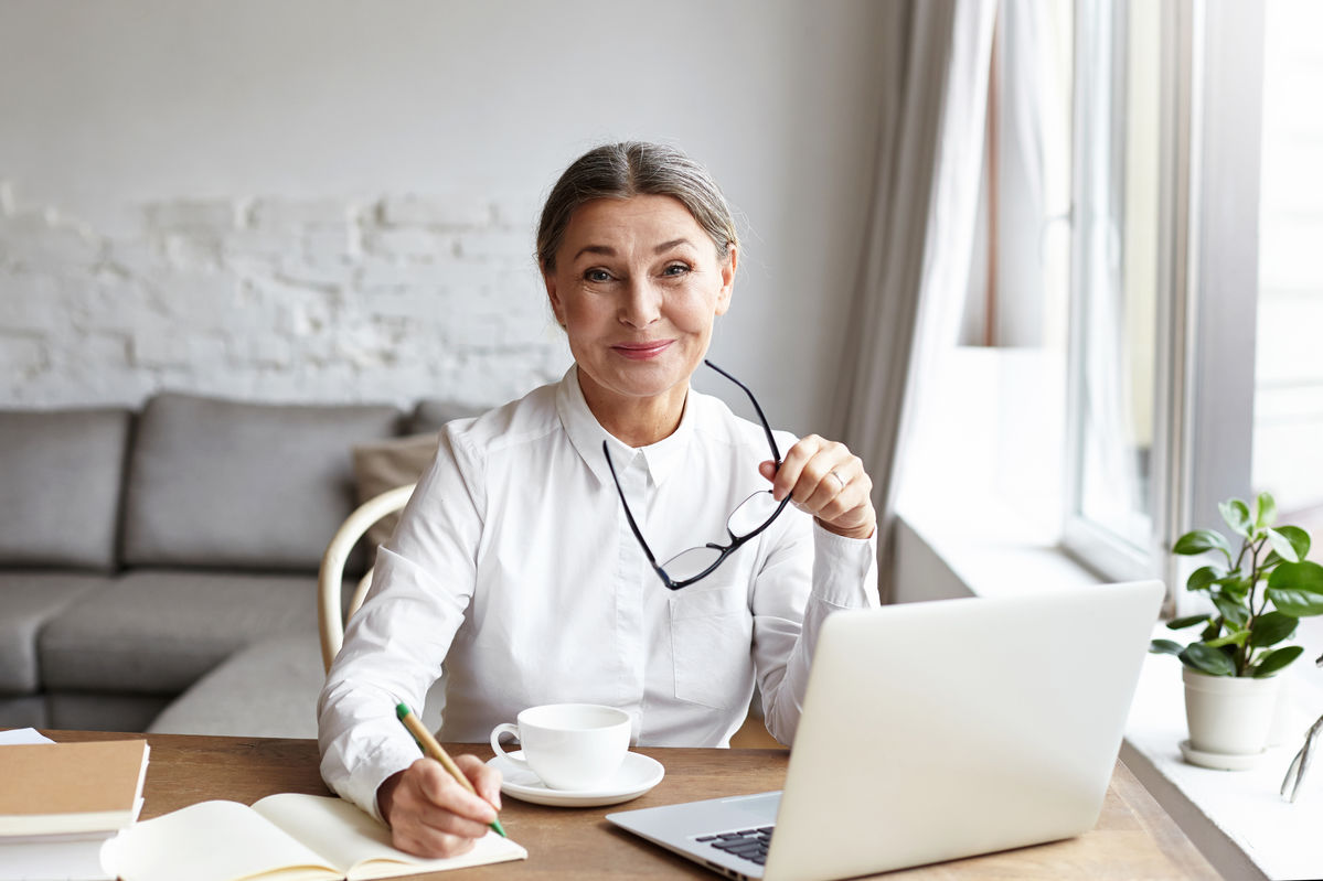 older woman sitting at desk with laptop holding glasses