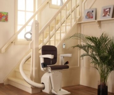 A curved stairlift ascends a wooden staircase in a light-colored, residential interior.  The lift is positioned near a potted plant and framed family photos.