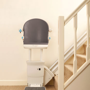 A stair lift sits folded against a carpeted staircase, ready to transport someone up the stairs.  The lift is mostly white and grey, with light blue accents.