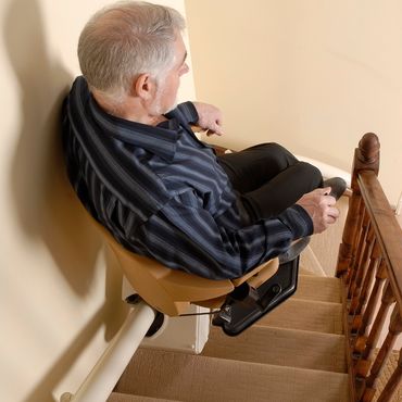 A man sits in a beige stairlift, ascending a carpeted staircase with a wooden banister; the lift is mounted to the wall.