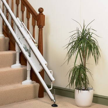 A stairlift, folded against a carpeted staircase, is situated in a home environment.  A potted plant stands nearby.