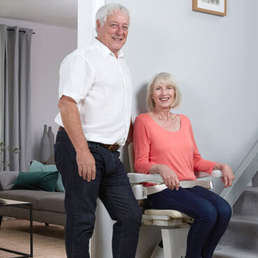 A seated woman and standing man smile near a stairlift inside a home.