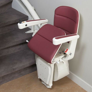 A burgundy-padded stair lift chair sits on a carpeted staircase,  partially folded up, ready to ascend.  The lift is attached to a curved track.