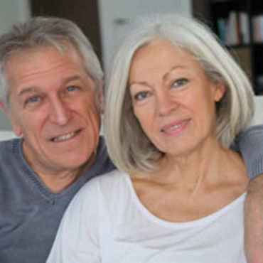 A senior couple sits close together, smiling gently at the camera, in what appears to be their living room.