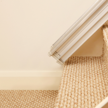 A light-grey stair lift is being installed on a carpeted staircase.  The lift's track is affixed to the wall and steps.