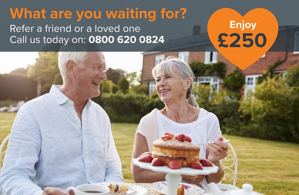A happy elderly couple enjoys a cake outdoors. They are seated at a table, sharing a strawberry cake in a garden.