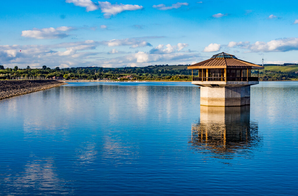 A hexagonal structure with a gazebo-like top sits on a calm lake.  It reflects in the still water, with a distant shoreline of trees and buildings visible in the background under a partly cloudy sky.